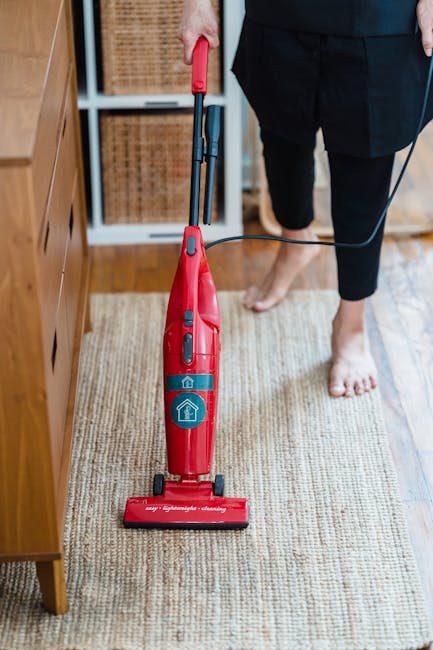 A close-up of a dry vacuum cleaner with a clear dust container and a black base, positioned on a pink carpeted floor in a residential room. The vacuum's flexible hose is attached to the machine and ex